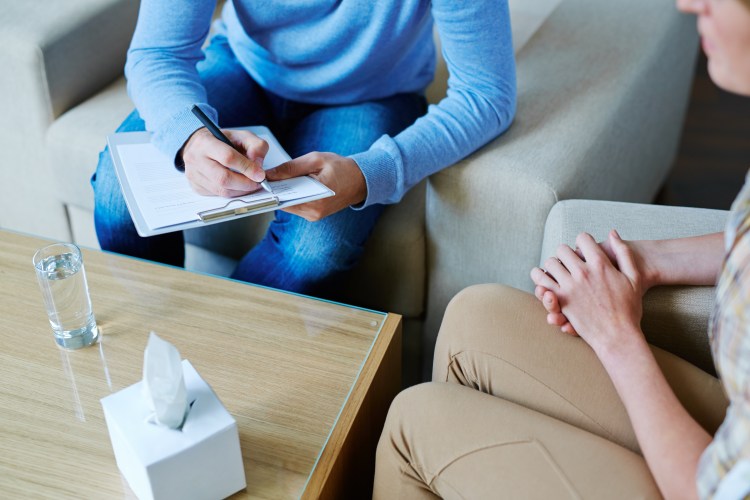 photo of a therapy session that only shows the hands of the individuals in the room as they are sitting on two sofa chairs. One person is taking notes.
